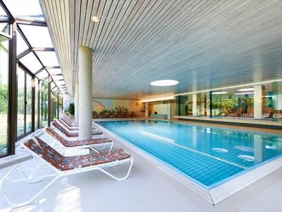 Piscina coperta moderna e luminosa con grandi finestre, lettini comodi e pilastri.