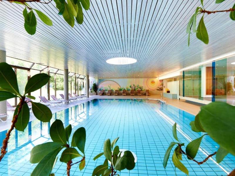 Piscina coperta luminosa con grandi finestre, piante in primo piano e soffitto moderno.