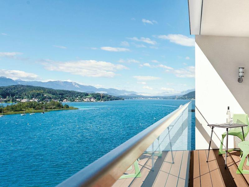 Balcone con ringhiera in vetro e sedie verdi con vista su un lago blu e montagne in una giornata di sole.