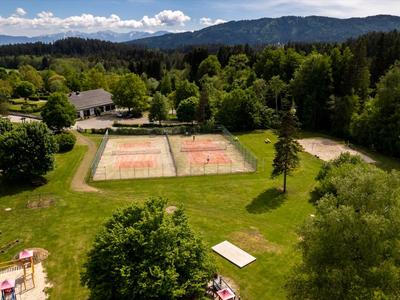 Vista aerea di un resort con due campi da tennis in sabbia circondati da alberi e montagne sullo sfondo.