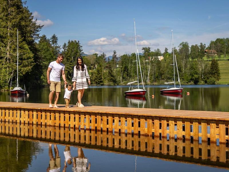 Een stel loopt op een houten steiger over een meer met zeilboten en bomen op de achtergrond.