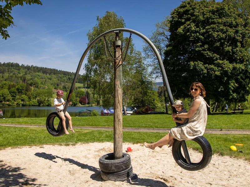 Twee personen schommelen aan bandenschommels in een zandige speeltuin in een zonnig park.