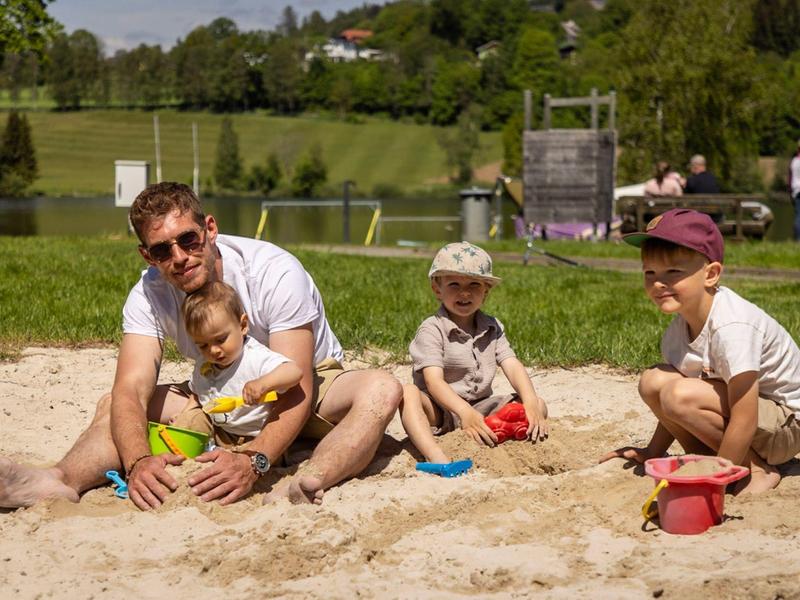 Vader en drie kinderen spelen samen met zand speelgoed in een zandbak buiten.