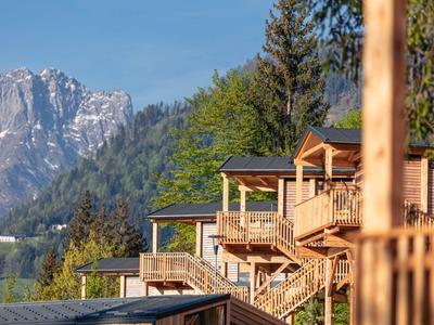 Cabañas de madera con escaleras frente a montañas boscosas y cielo azul.
