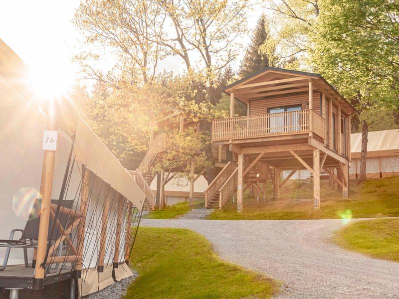 Sonniger Campingplatz mit Zelt und erhöhtem Holzhäuschen umgeben von Bäumen und Wiese.