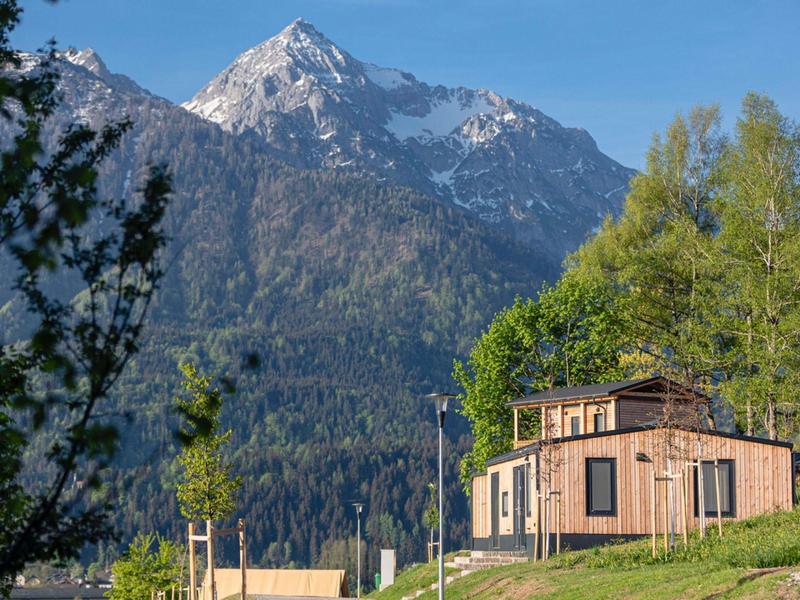 Moderner Holzbungalow vor grünen Bäumen mit schneebedecktem Berg und blauem Himmel im Hintergrund.