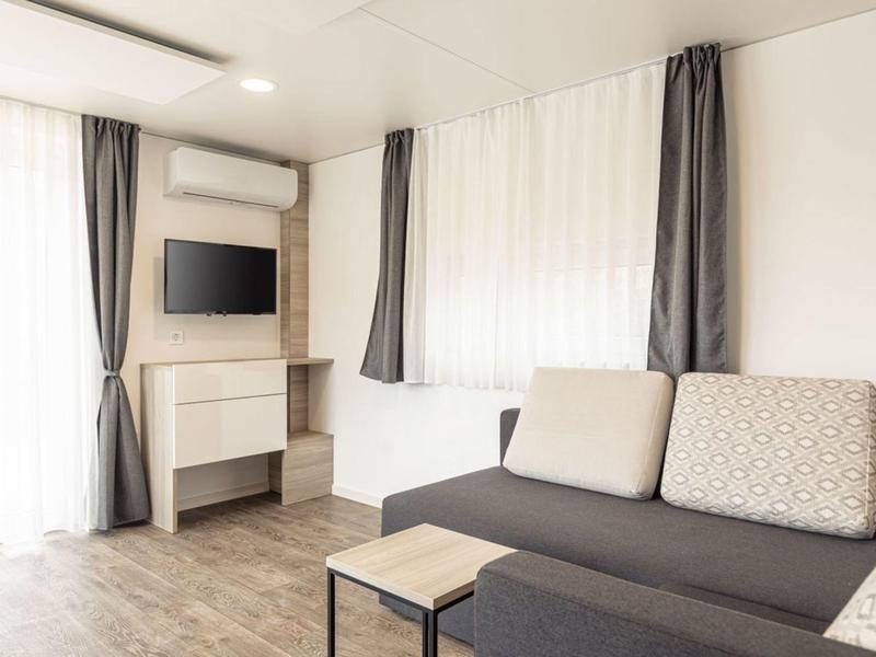 Helles Wohnzimmer mit grauer Couch, kleinem Tisch, Flachbildschirm, weißen Wänden und Vorhängen.