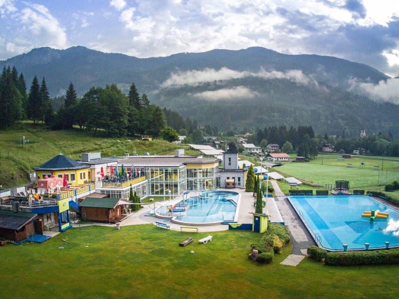 Bergresort mit Schwimmbädern, grünen Wiesen und bewaldeten Hügeln unter blauem Himmel.
