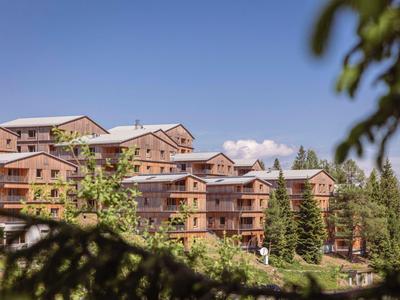 Mehrere große Holzhäuser mit Balkonen stehen in einer grünen Berglandschaft unter blauem Himmel.