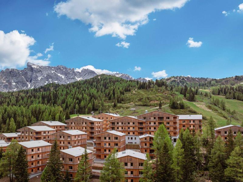 Mehrere Holzhäuser stehen auf einer grünen Bergwiese vor bewaldeten Hügeln und Bergen unter blauem Himmel.