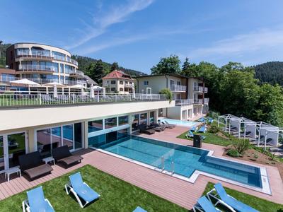 Moderner Hotelkomplex mit Pool, Liegestühlen und grünen Hügeln im Hintergrund.