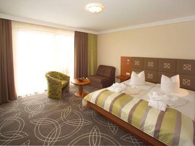 Helles Hotelzimmer mit Doppelbett, Sesseln, Holzmöbeln und großem Fenster mit Vorhängen.