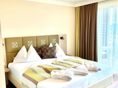 Helles Hotelzimmer mit Doppelbett, bunten Kissen und großen Fenstern mit Vorhängen.
