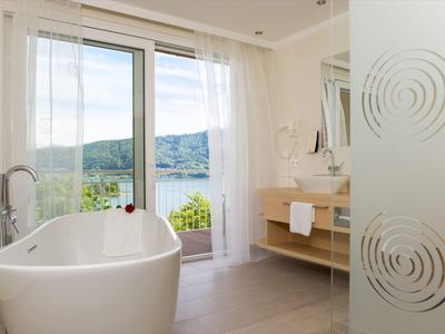 Helles Badezimmer mit freistehender Badewanne und großer Glasfront mit Blick auf See und Hügel.