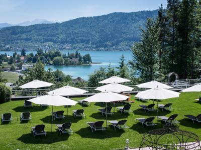 Grüne Wiese mit Liegestühlen und weißen Sonnenschirmen vor Bergsee und bewaldeten Hügeln.