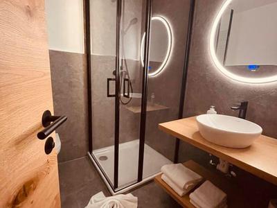 Nowoczesna łazienka z okrągłym podświetlanym lustrem, szklaną kabiną prysznicową i drewnianą półką z umywalką.