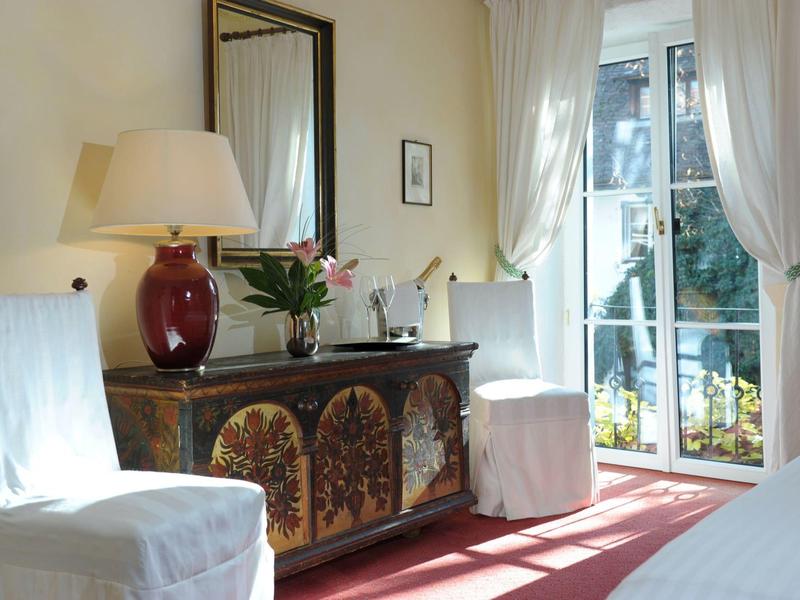 Hell eingerichtetes Zimmer mit weiß bezogenen Stühlen, Kommode, Lampe und großen Fenstern.