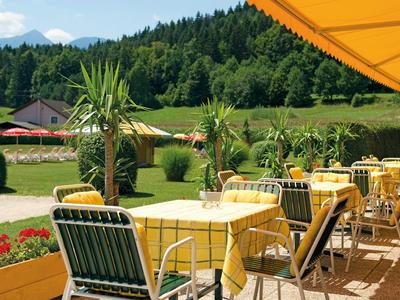 Sonnige Terrasse mit gelben Tischen und Stühlen, grünen Pflanzen und Bergen im Hintergrund.