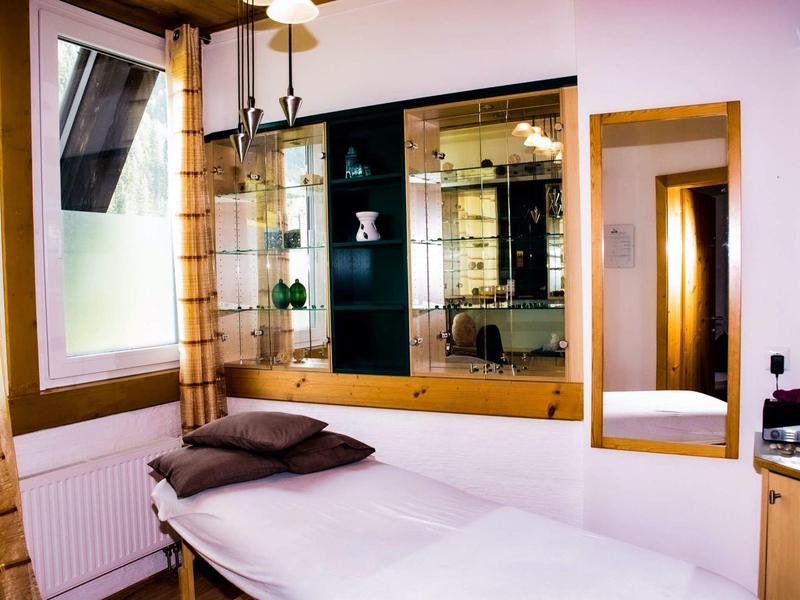 Jasny pokój z łóżkiem do masażu, lustrem i oknem z zasłonami.