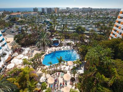 Vista de una piscina de hotel con tumbonas y palmeras alrededor, ciudad y mar al fondo.