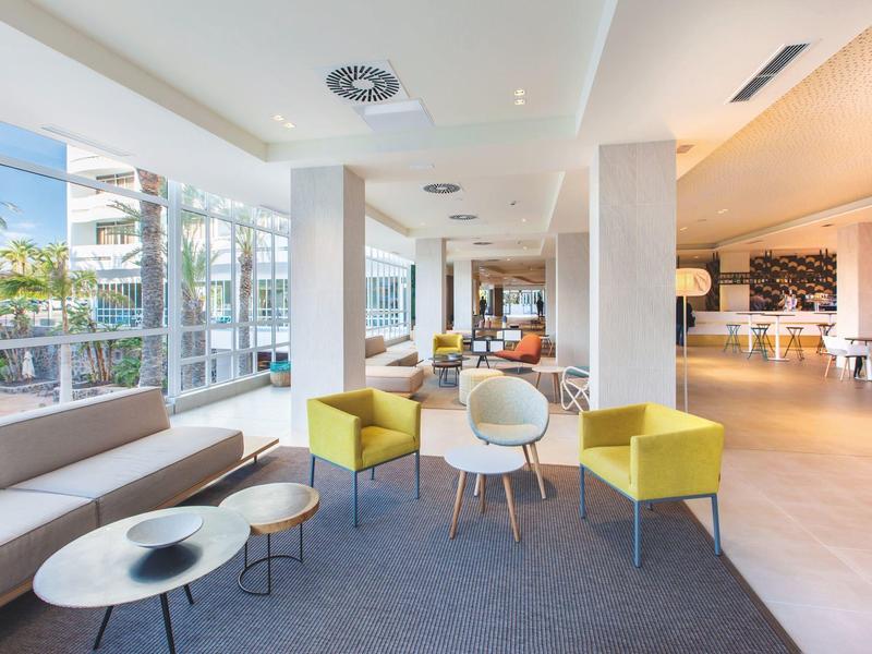 Sala moderna de hotel con sofás, sillas y grandes ventanales con vista a palmeras.