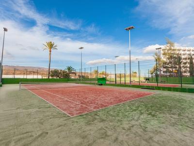 Freiluft-Tennisplatz mit Zaun und einem Hotelgebäude im Hintergrund unter blauem Himmel.