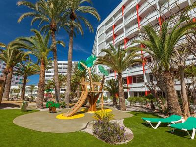 Jardin d'hôtel moderne avec palmiers, toboggan de terrain de jeu et chaises longues sous un ciel bleu.