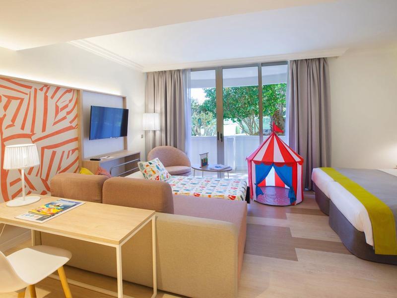 Chambre d'hôtel lumineuse avec coin salon, tente de jeu et grande fenêtre donnant sur des arbres.