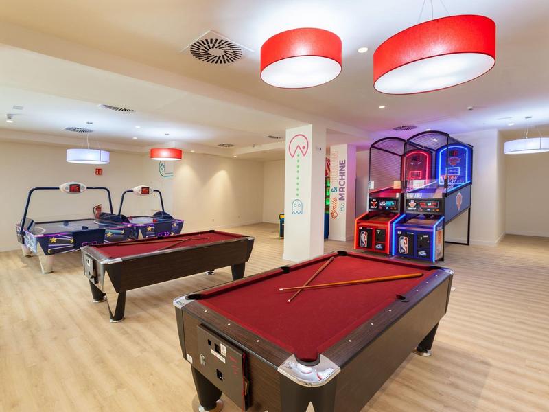 Salle de jeux avec trois tables de billard, deux tables de air hockey et plusieurs jeux d'arcade dans un espace lumineux.