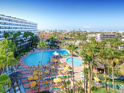 Hotel with large pool, sun loungers, umbrellas, and surrounding palm trees under clear sky.