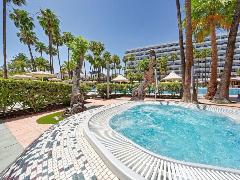 Outdoor hot tub with palm trees and a modern hotel in the background under blue sky.