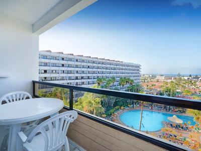 Balcony with white chairs and table overlooks pool, hotel, and distant city under blue sky.