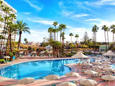 Großer Hotelpoolbereich mit Palmen, Sonnenliegen und blauem Himmel.