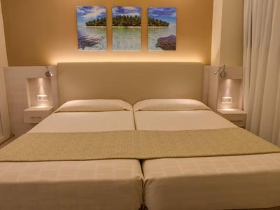 Twin beds with beige bedding in a warmly lit hotel room with art above the headboard.