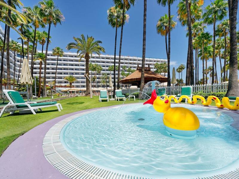 Hotel pool area with palm trees, lounge chairs, and a large yellow duck float.