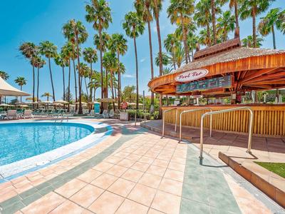 Outdoor pool with palm trees and a tiki bar under a clear blue sky at a resort.