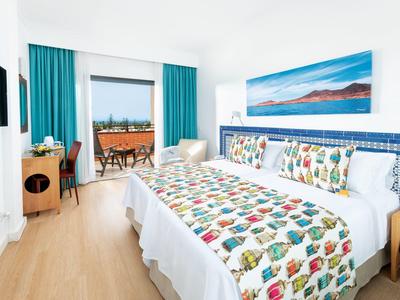 Habitación de hotel luminosa con dos camas individuales, colchas coloridas y vista al balcón con vista al mar.
