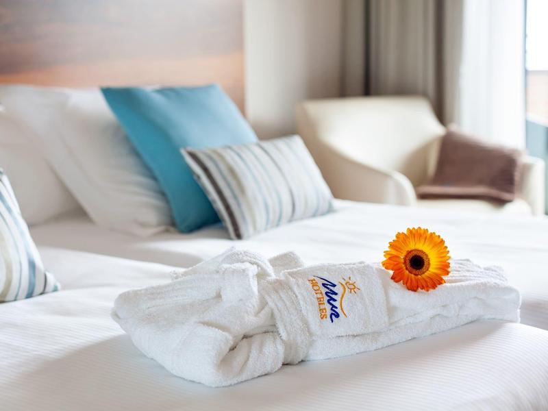 Una cama de hotel con toallas blancas, una margarita naranja y cojines en tonos azules y grises.