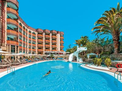 Curved hotel with a large pool, waterslide, palm trees, and sun loungers under a clear sky.