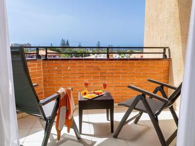 Small balcony with two chairs, a table, and a colorful towel, overlooking rooftops and sea.