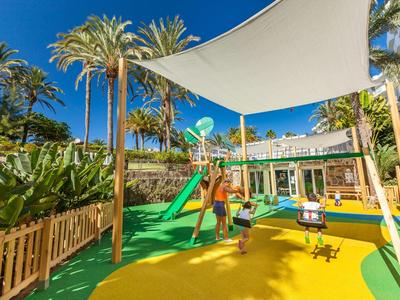 Bambini giocano in un parco giochi colorato con scivolo sotto una tenda da sole e palme.