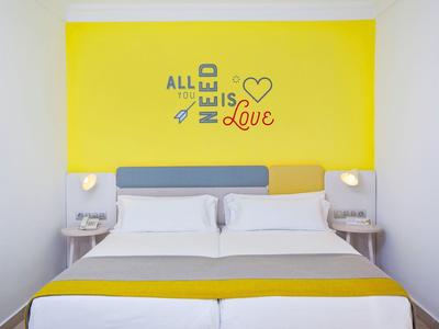 Camera d'hotel moderna con due letti singoli, accenti gialli e decorazione murale con citazione sull'amore.