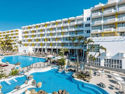 Großes Hotel mit mehreren Stockwerken, Swimmingpools und Palmen unter klarem blauem Himmel.