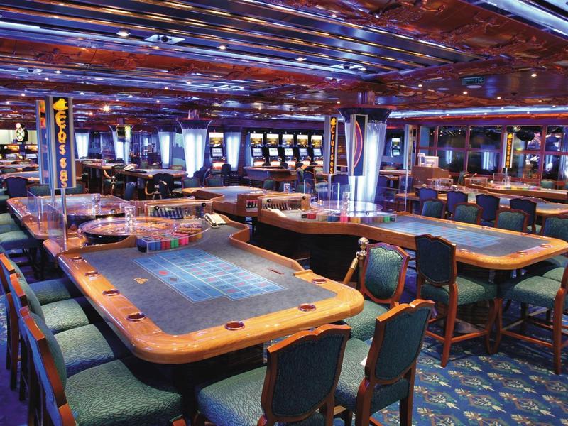 Casino gaming tables with green chairs under a patterned ceiling and blue carpet.