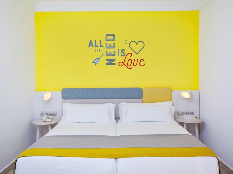 Modern hotel room with two single beds, yellow accents, and a wall decoration with a love quote.