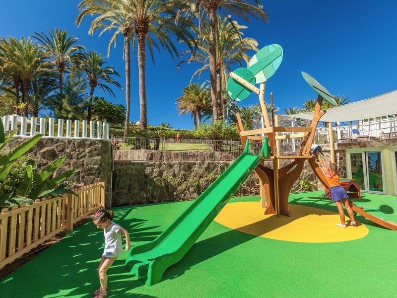 Children playing on a colorful playground with slide and climbing frame under palm trees.