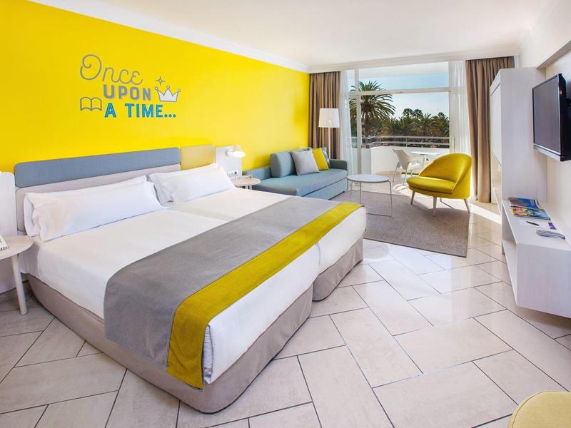 Bright hotel room with large bed, yellow accent wall, seating area, and balcony overlooking palm trees.