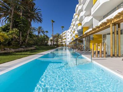 Modern hotel complex with a long pool and palm trees in a sunny setting.