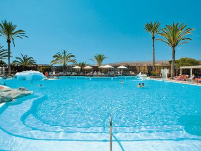 Large blue hotel pool with palm trees, lounge chairs, and clear sky in the background.