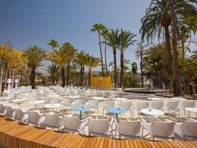 Modern outdoor area with white seating and blue tables under palm trees in clear sky.